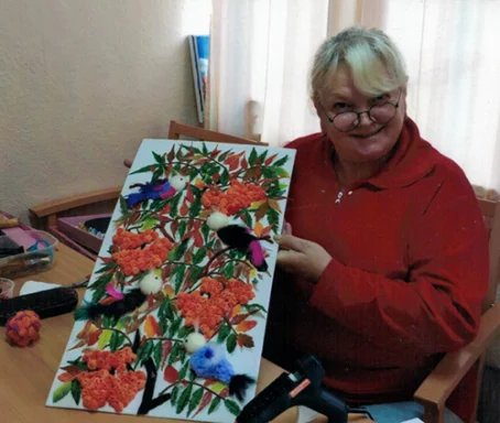 Kunst in der TAL Frau mit roten Pullover hält ein farbenfrohes Kunstwerk mit Vögeln und Blumen.
